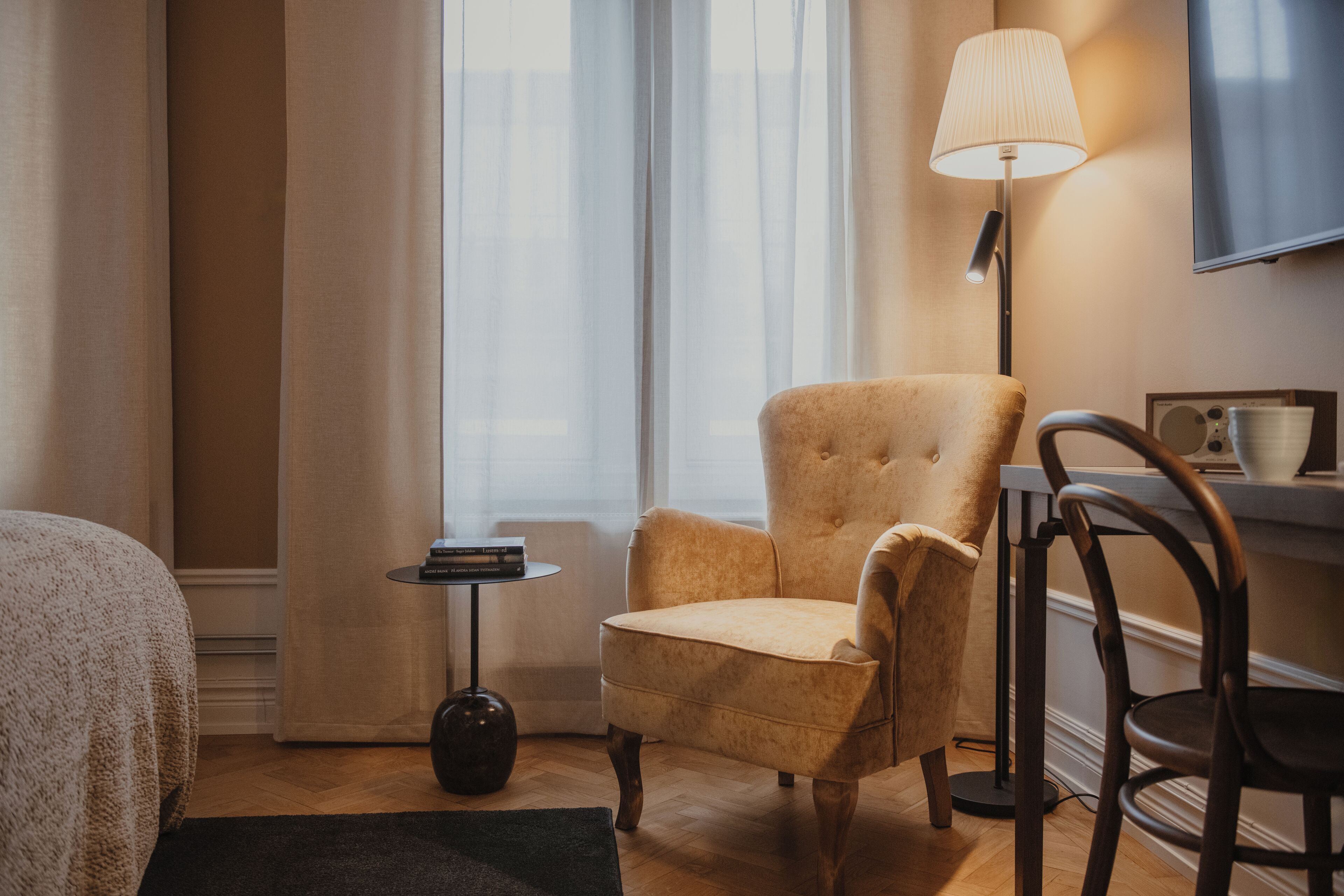 Habitación de hotel con un ventanal grande, un sofá, una silla, una mesita auxiliar, una lámpara y un televisor de pantalla plana montado en la pared.