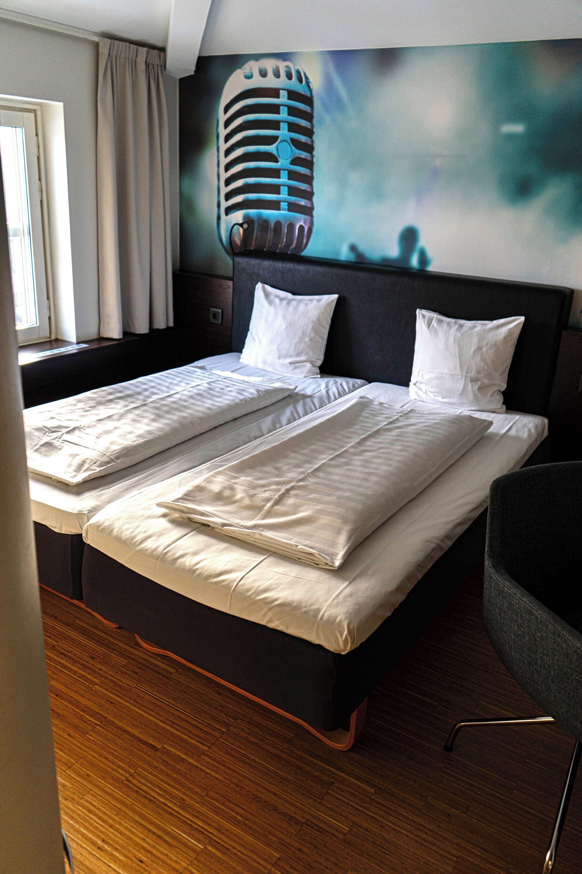 A hotel room with a large bed, a headboard with a microphone design, and a window with curtains.