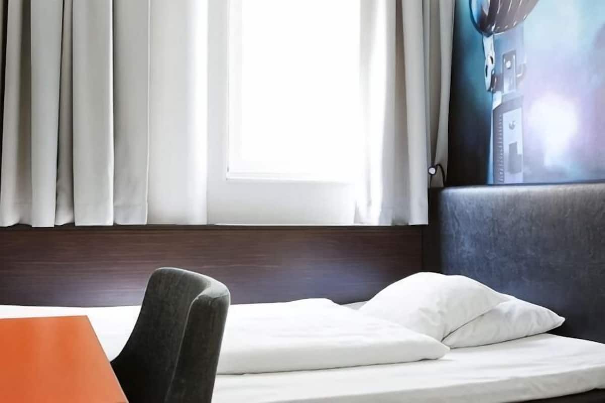 Una habitación de hotel con cama, una silla, un ventanal con cortinas y una lámpara fijada a la pared.