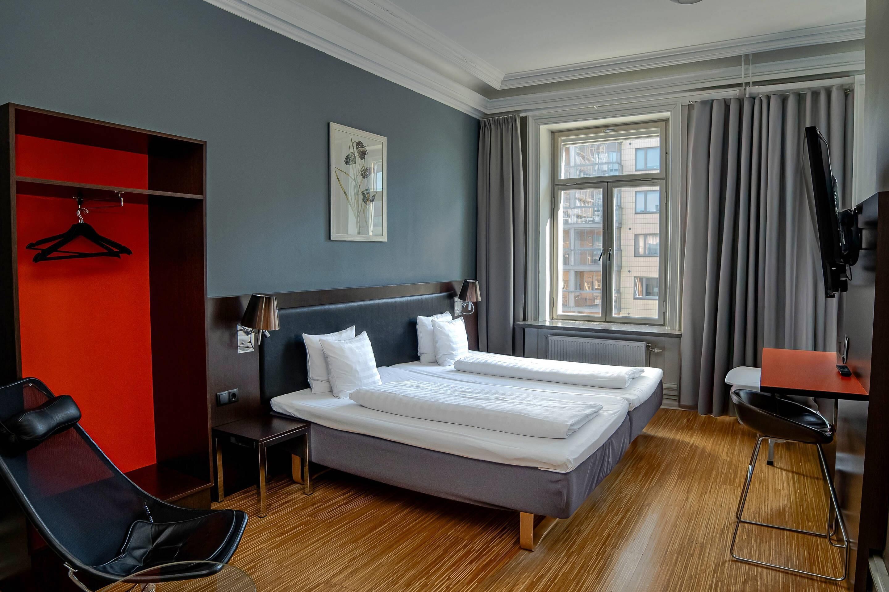 A hotel room with a bed, a chair, a desk, a TV, a window with curtains, and a red wall.