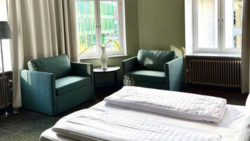 A hotel room with a bed, two green armchairs, a small table, and a window with curtains.