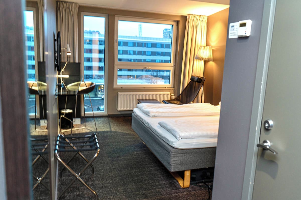 Habitación de hotel con cama, dos sillas, escritorio, lámpara y vista a un edificio a través del ventanal.