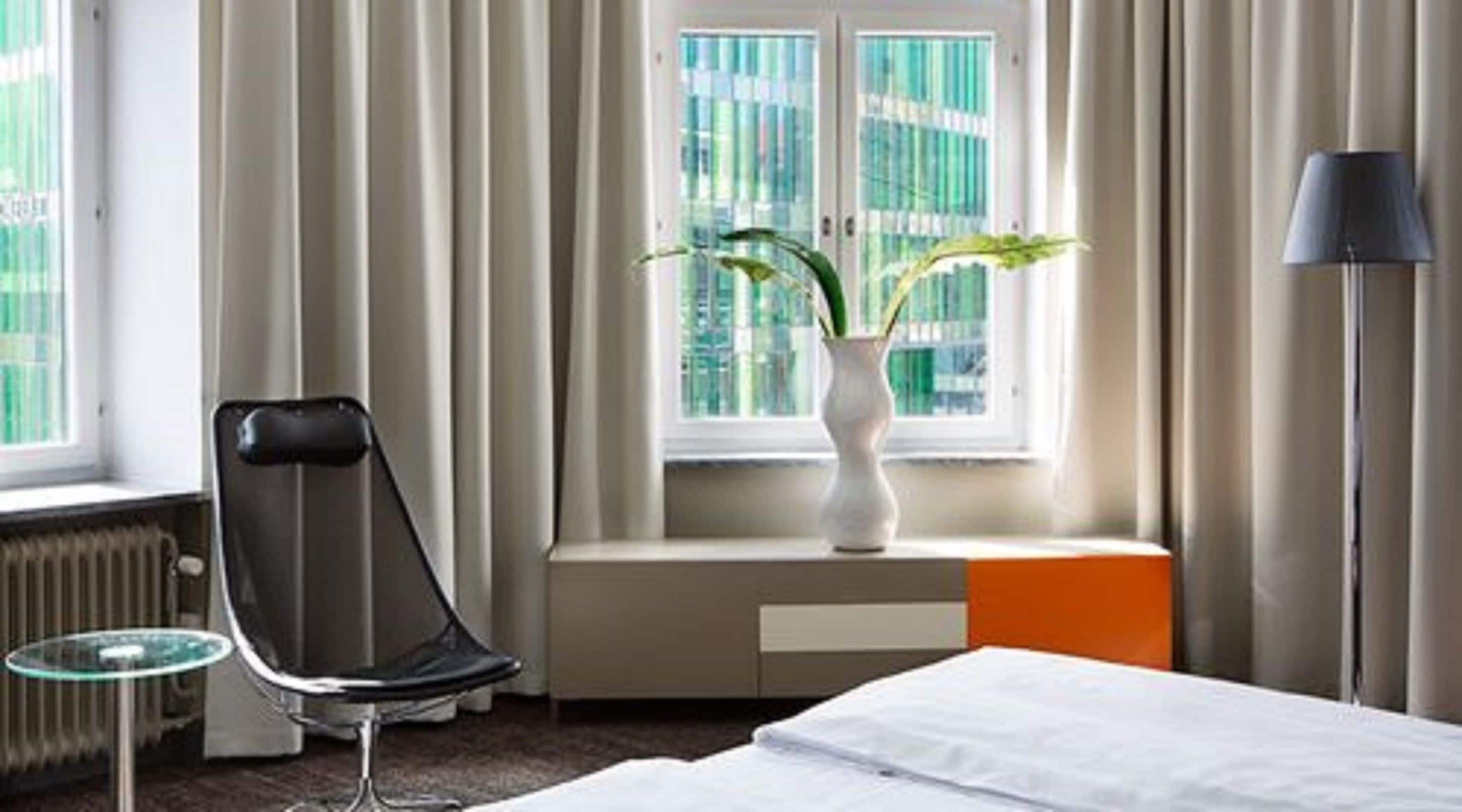 Een hotelkamer met een bed, een stoel, een kleine tafel, een vaas met planten en een raam met gordijnen.