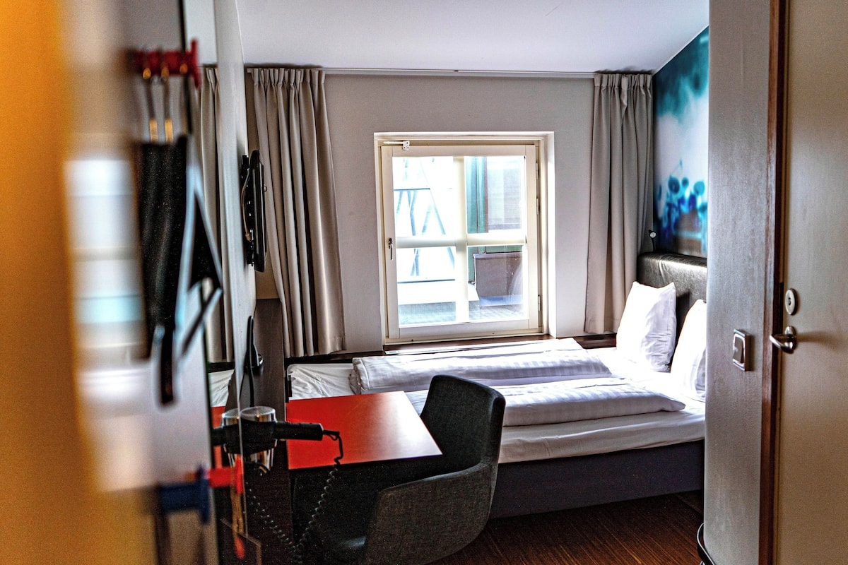 Habitación de hotel con cama, escritorio, silla, televisión y ventana con cortinas.