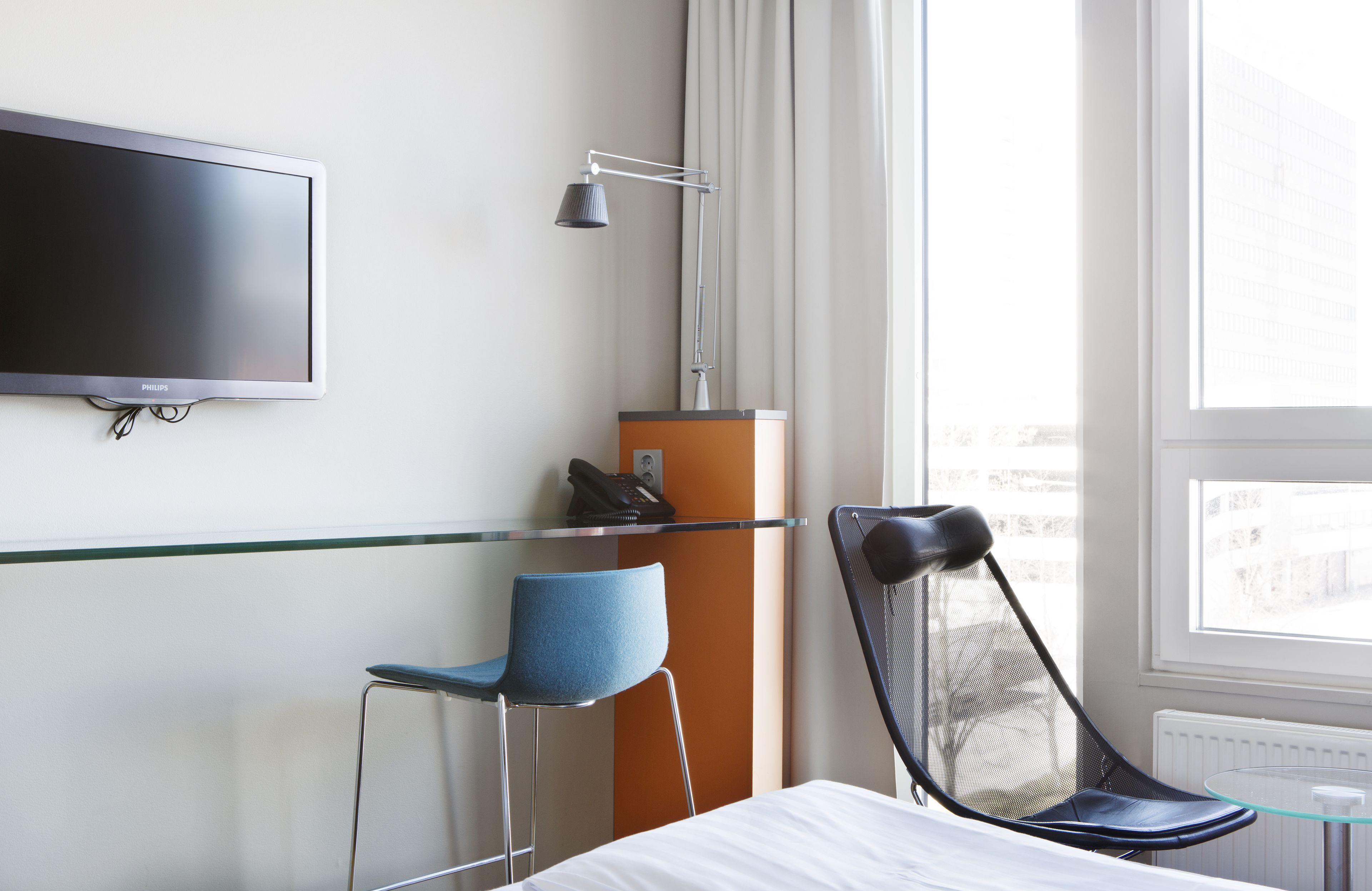 A hotel room with a bed, a TV mounted on the wall, a desk with a chair, a standing lamp, and a window with a view of buildings.