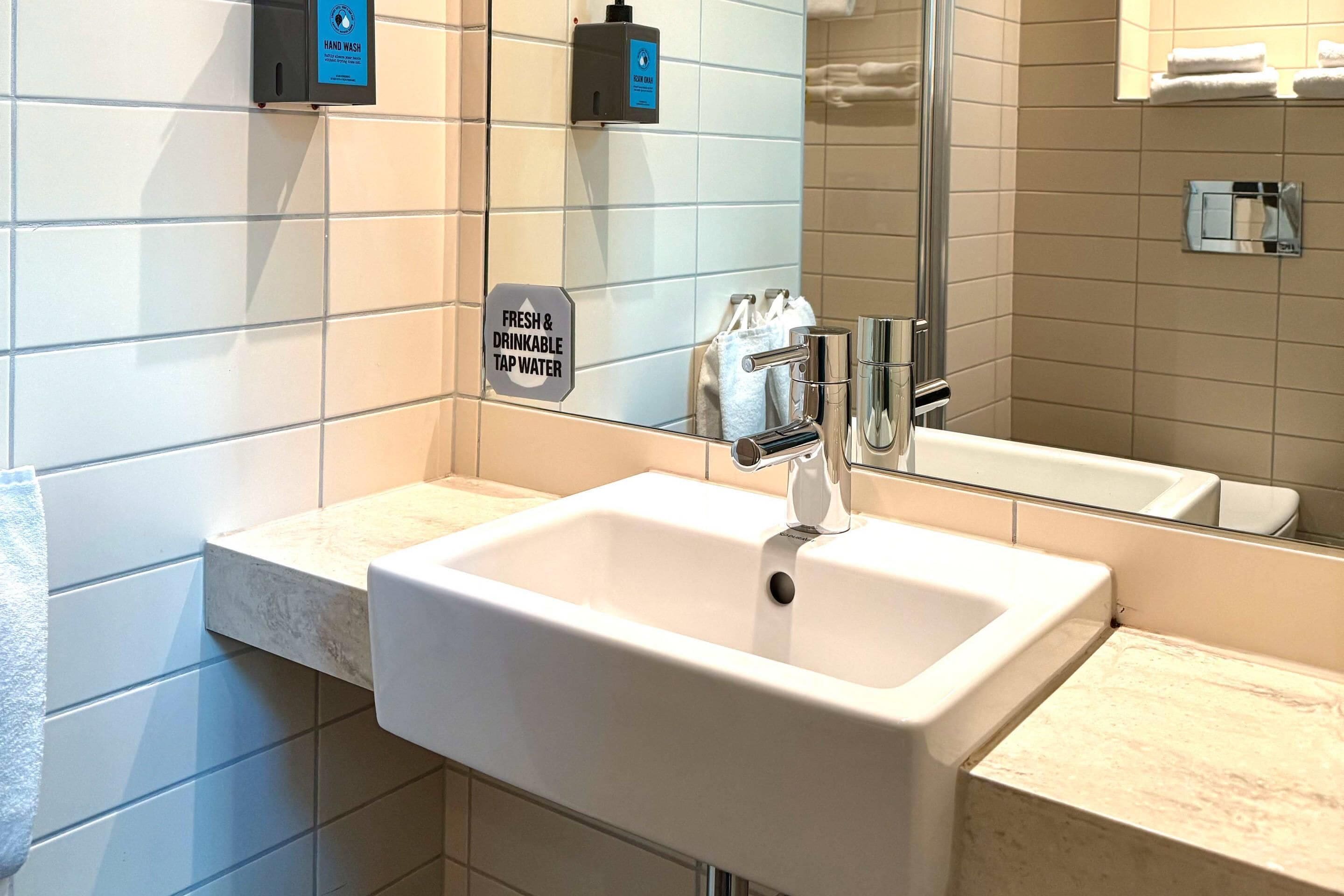 Een badkamer met een witte wasbak, zeepdispenser en een bordje dat vers en drinkbaar kraanwater aangeeft.