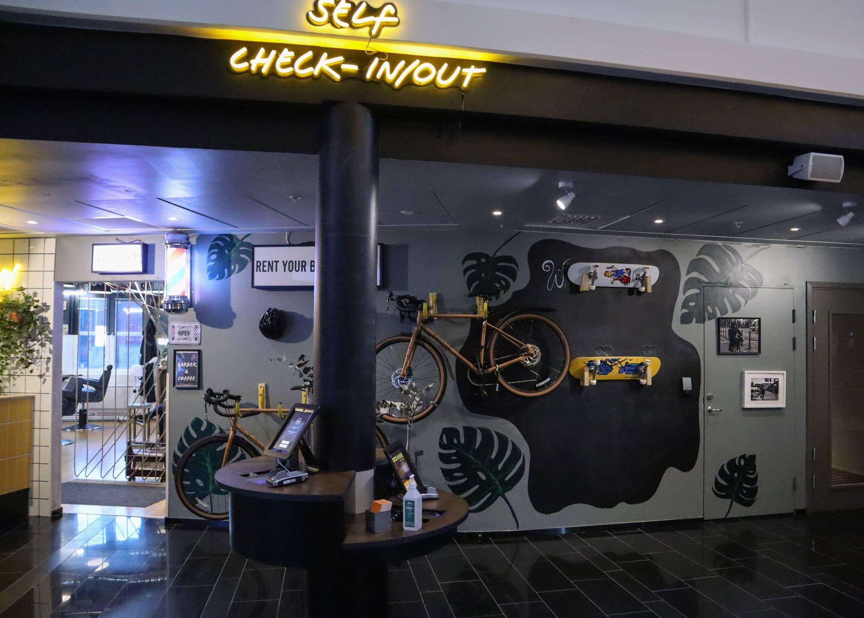 A self-check-in/out area with a neon sign, a bicycle, and a wall with plant and leaf designs.