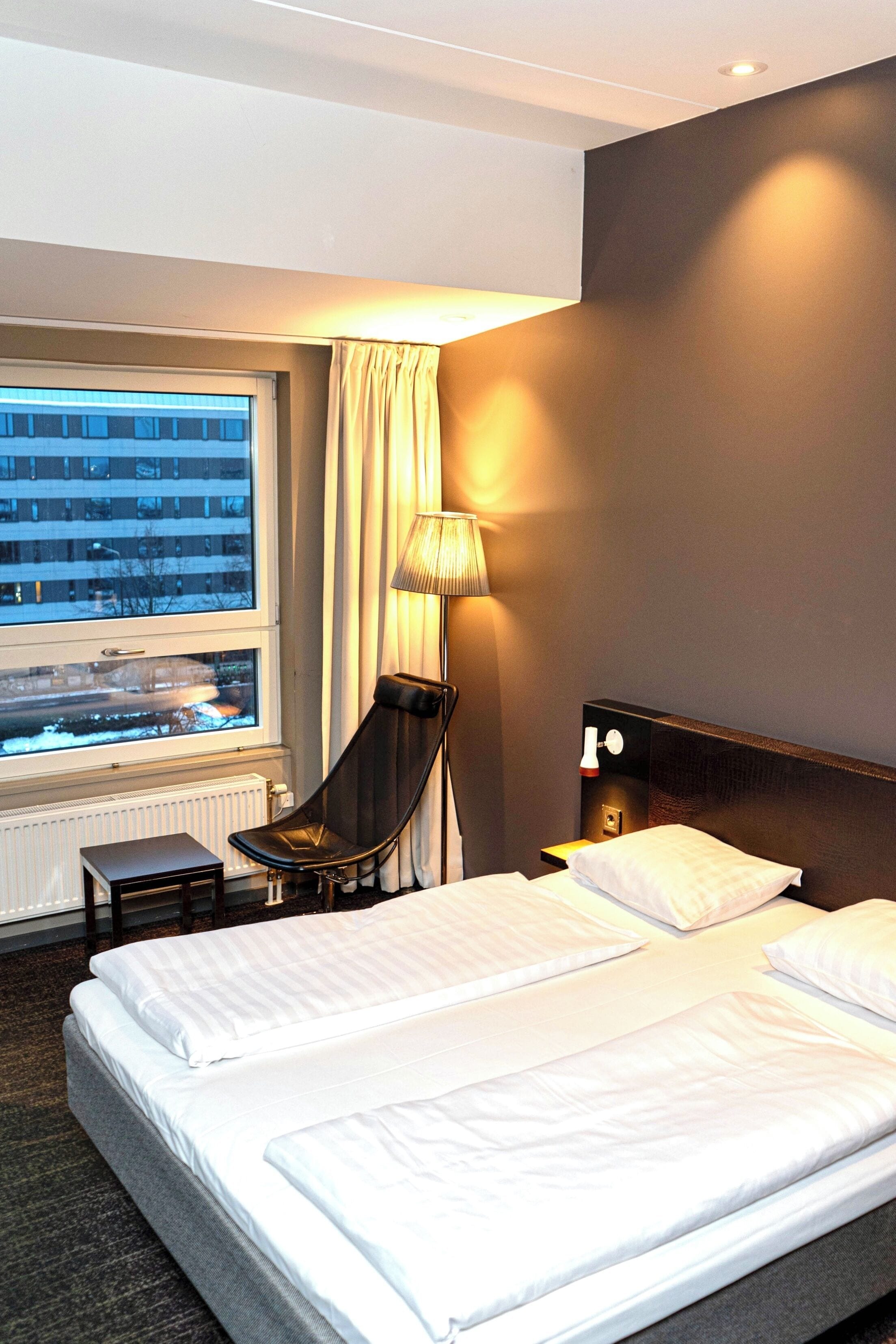 Une chambre d’hôtel avec un lit, une chaise, une lampe, une fenêtre donnant sur des immeubles et un radiateur.