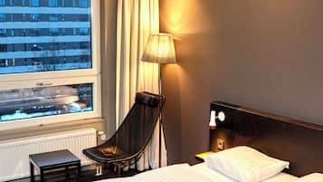 Une chambre d’hôtel avec un lit, une chaise, une lampe, une fenêtre donnant sur des immeubles et un radiateur.