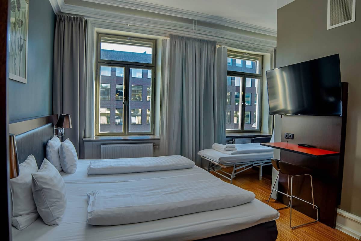 Habitación de hotel con dos camas, televisión, escritorio y ventanas grandes con cortinas.