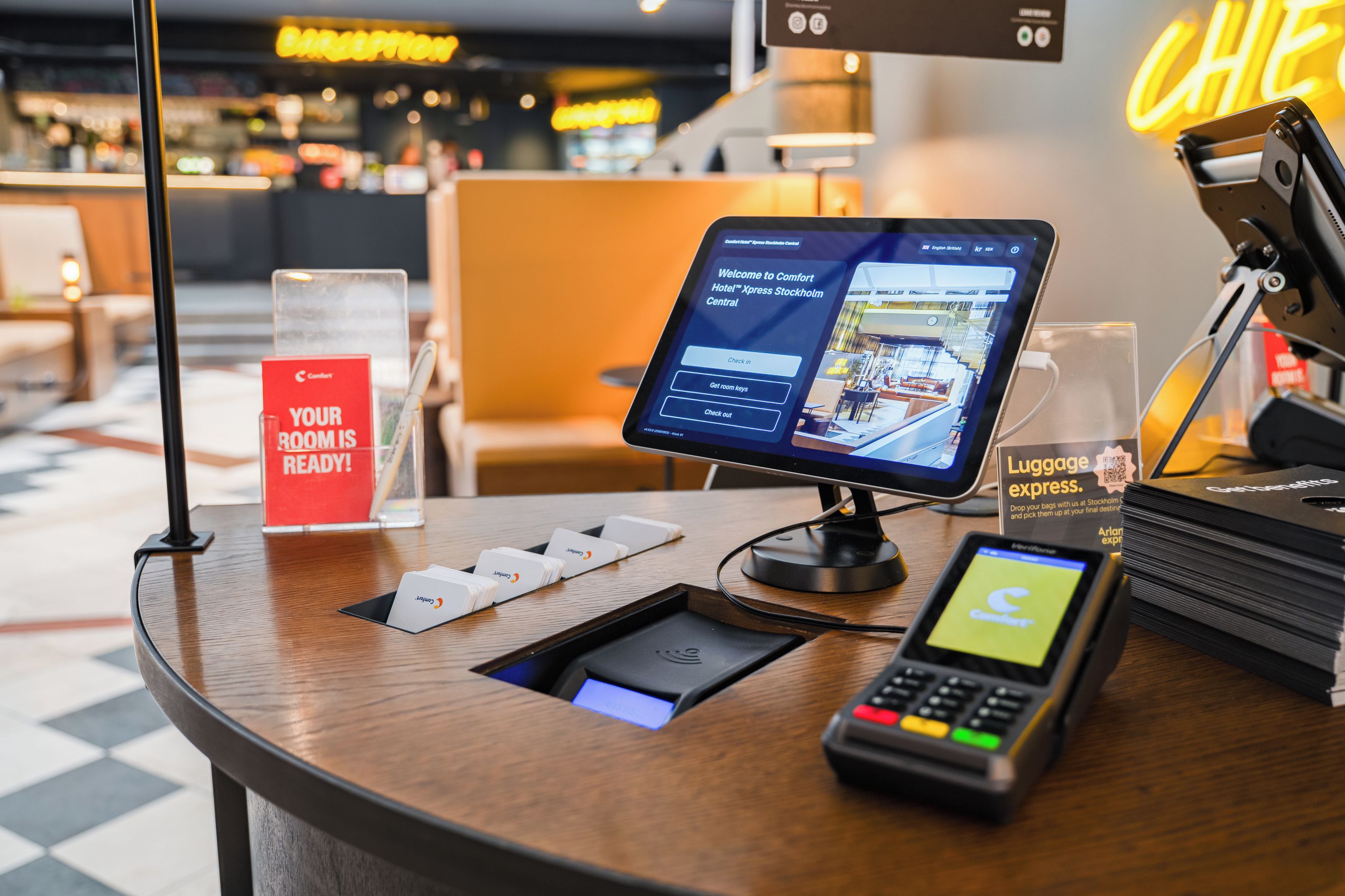 Un mostrador de recepción de hotel con un quiosco digital, un letrero de entrega rápida de equipaje y un aviso que indica que las habitaciones están listas.