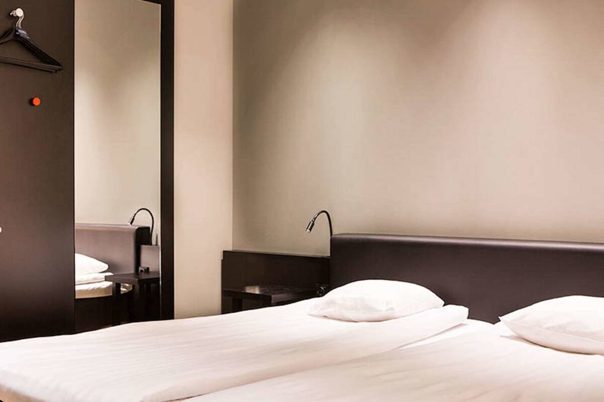 A hotel room with a bed, a bedside table, a mirror, and a wall-mounted light fixture.