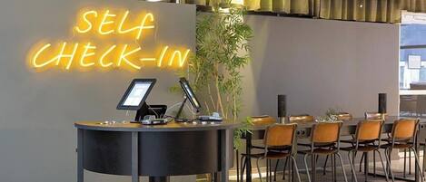 A self-check-in area with a neon sign, a computer, and a plant.