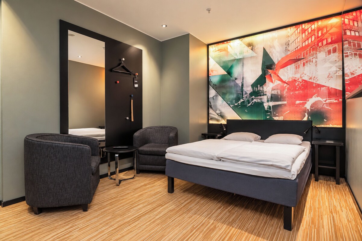Een moderne hotelkamer met een groot bed, twee fauteuils, een bureau en een spiegel.