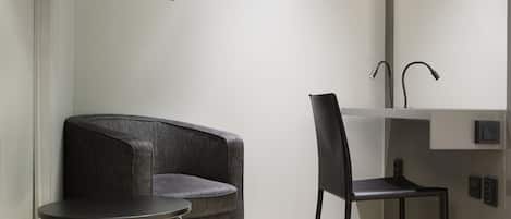 Una habitación moderna con un sofá gris, una mesita redonda pequeña, una silla negra y un lavamanos empotrado con espejo.