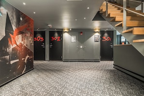 A hotel lobby with a modern design, featuring a staircase, multiple doors with room numbers, and a large abstract artwork on the wall.