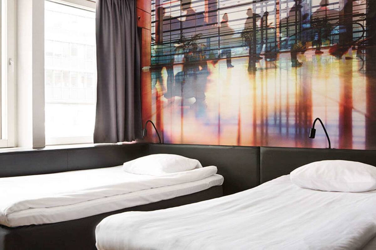 A hotel room with two single beds, a large window with curtains, and a colorful abstract wall art.