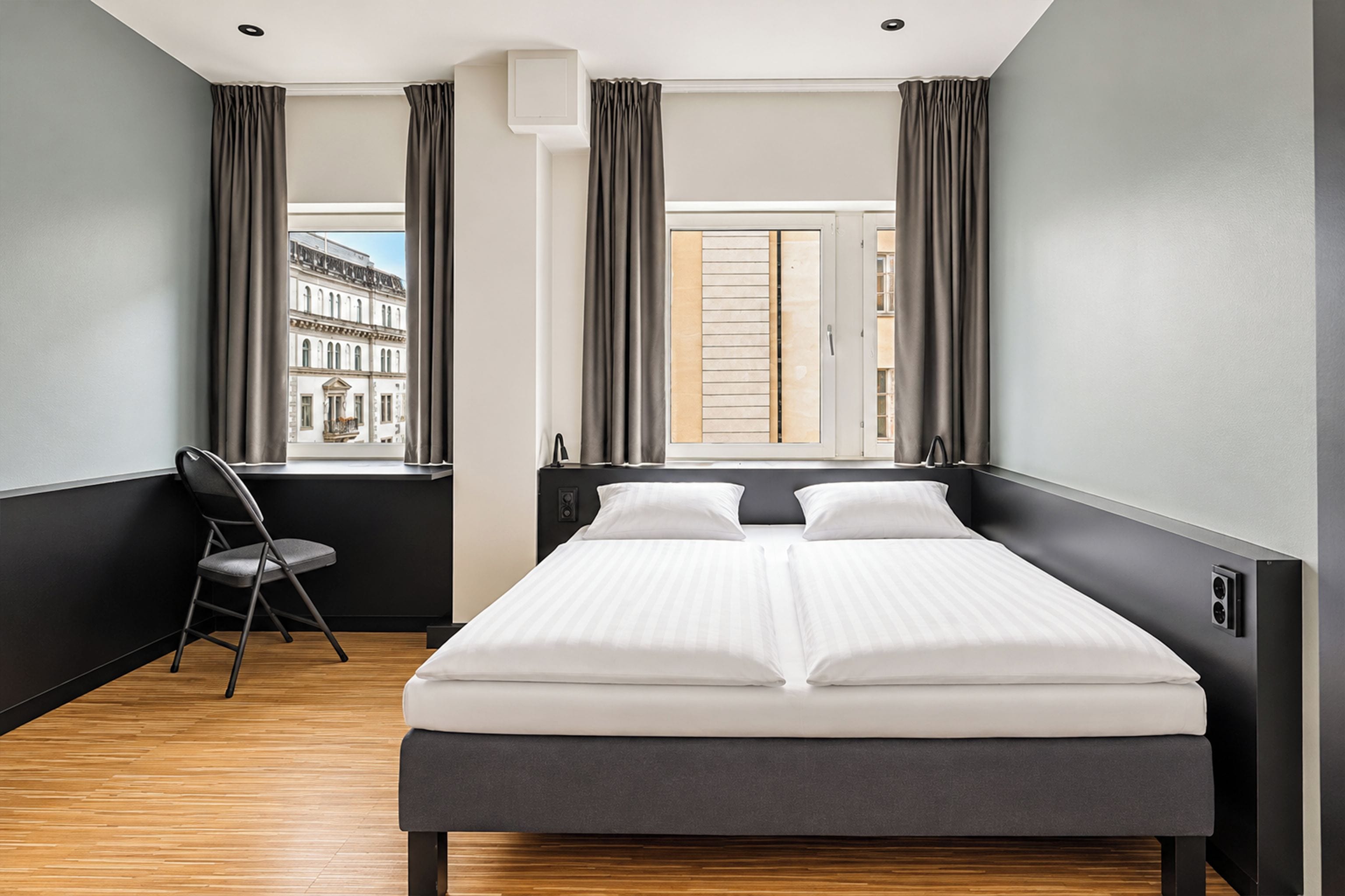 A hotel room with a bed, a chair, a window with curtains, and a view of a building outside.