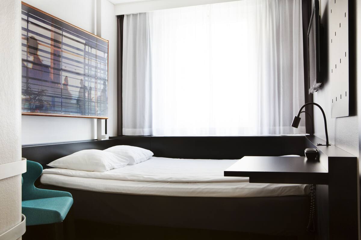 A modern hotel room with a single bed, a desk with a lamp, a view of the city through large windows, and a painting on the wall.