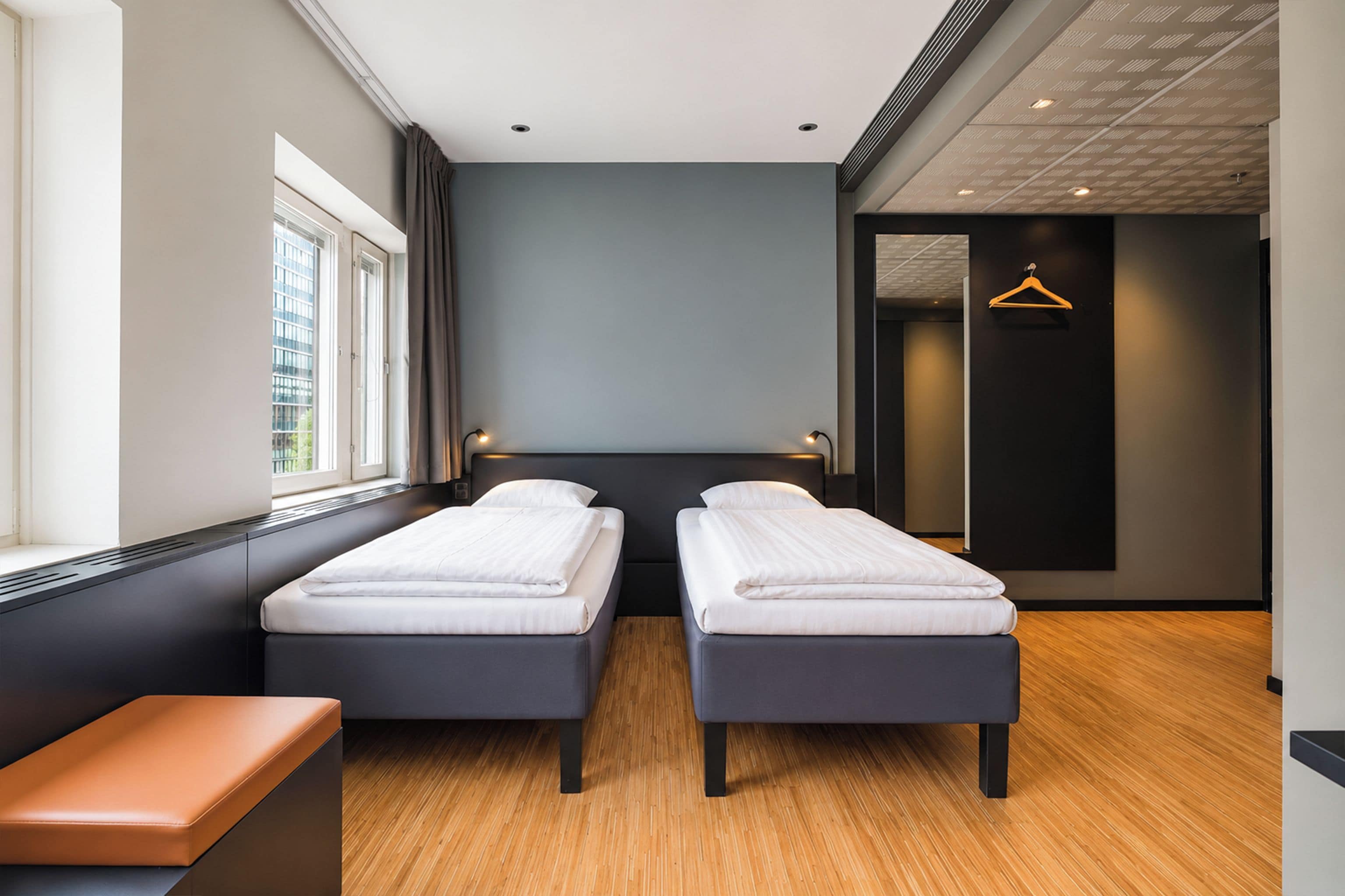 A modern hotel room with two single beds, a bench, a window, and a door leading to a hallway.