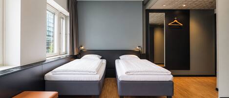 A modern hotel room with two single beds, a bench, a window, and a door leading to a hallway.