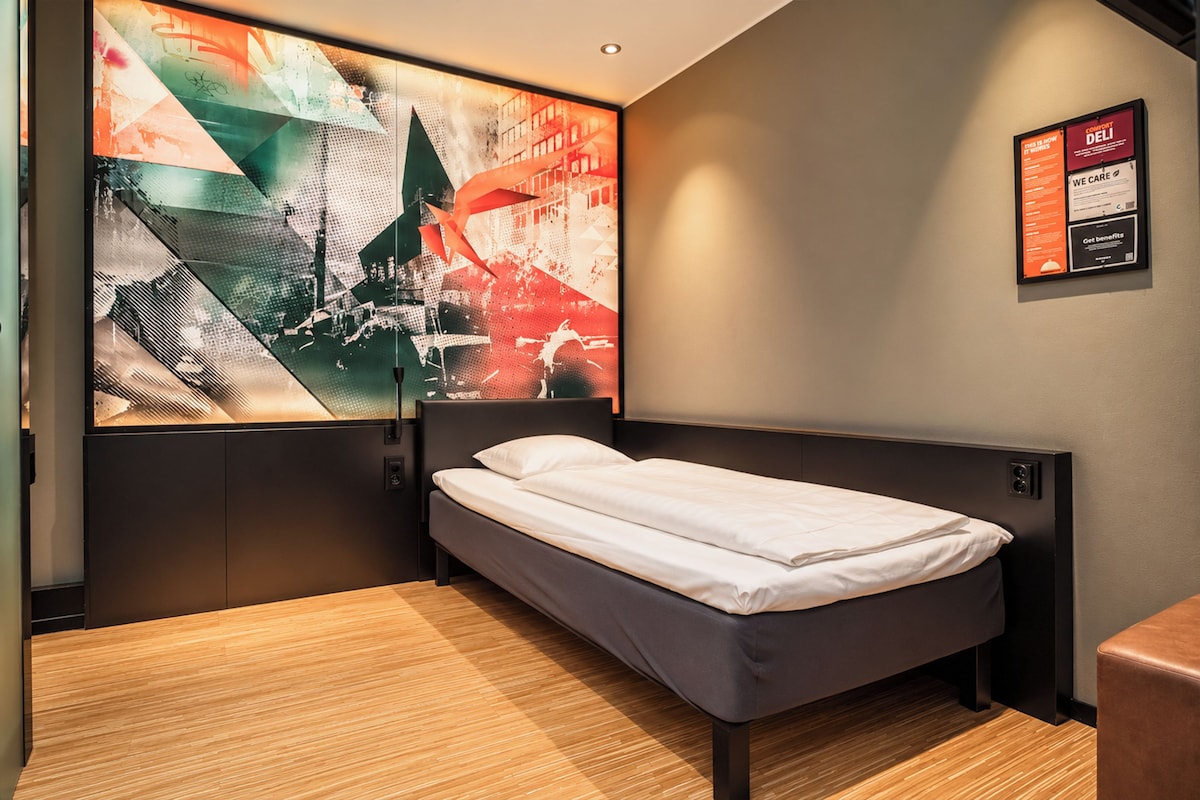 Een hotelkamer met een enkel bed, een groot abstract kunstwerk aan de muur en een ingelijst menu.