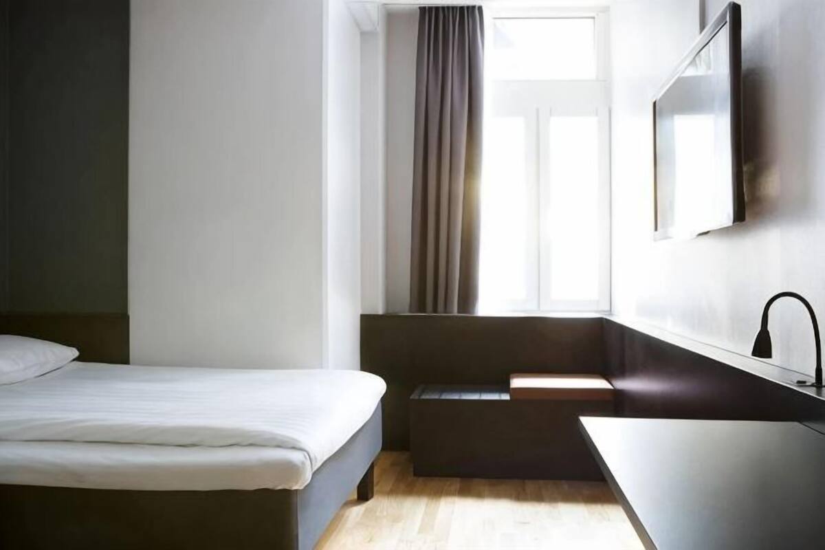 A hotel room with a bed, a small seating area, a window with curtains, a sink, and a mirror.
