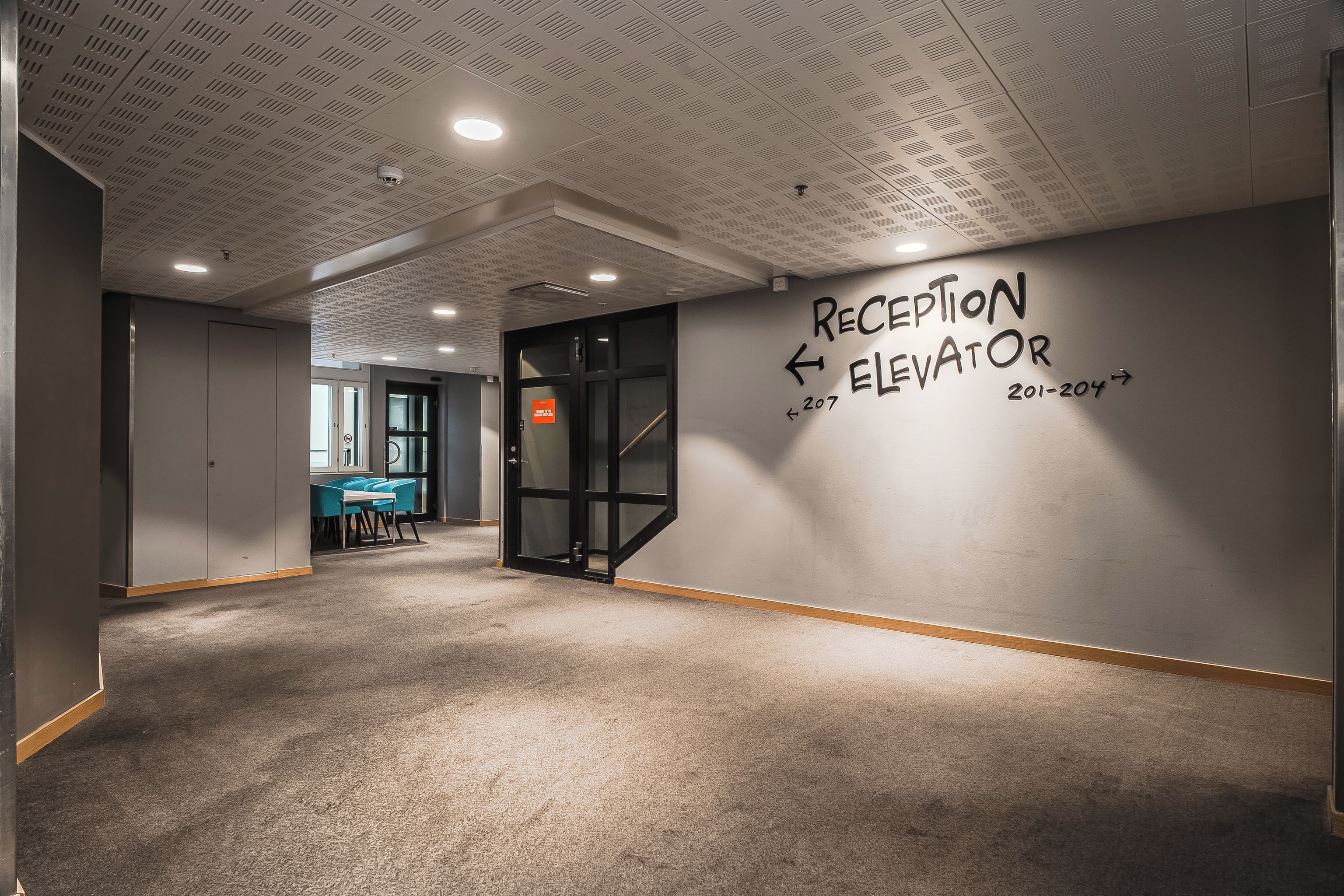 Een receptiegedeelte met een bord dat de richting van de lift aangeeft en een deur naar een ruimte met een bureau en stoelen.