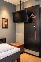 Una habitación de hotel con una cama, un televisor montado en la pared, un perchero de madera y un taburete pequeño de madera.