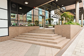 A modern building entrance with a set of stairs leading to the entrance, a glass facade, and a landscaped area with plants.
