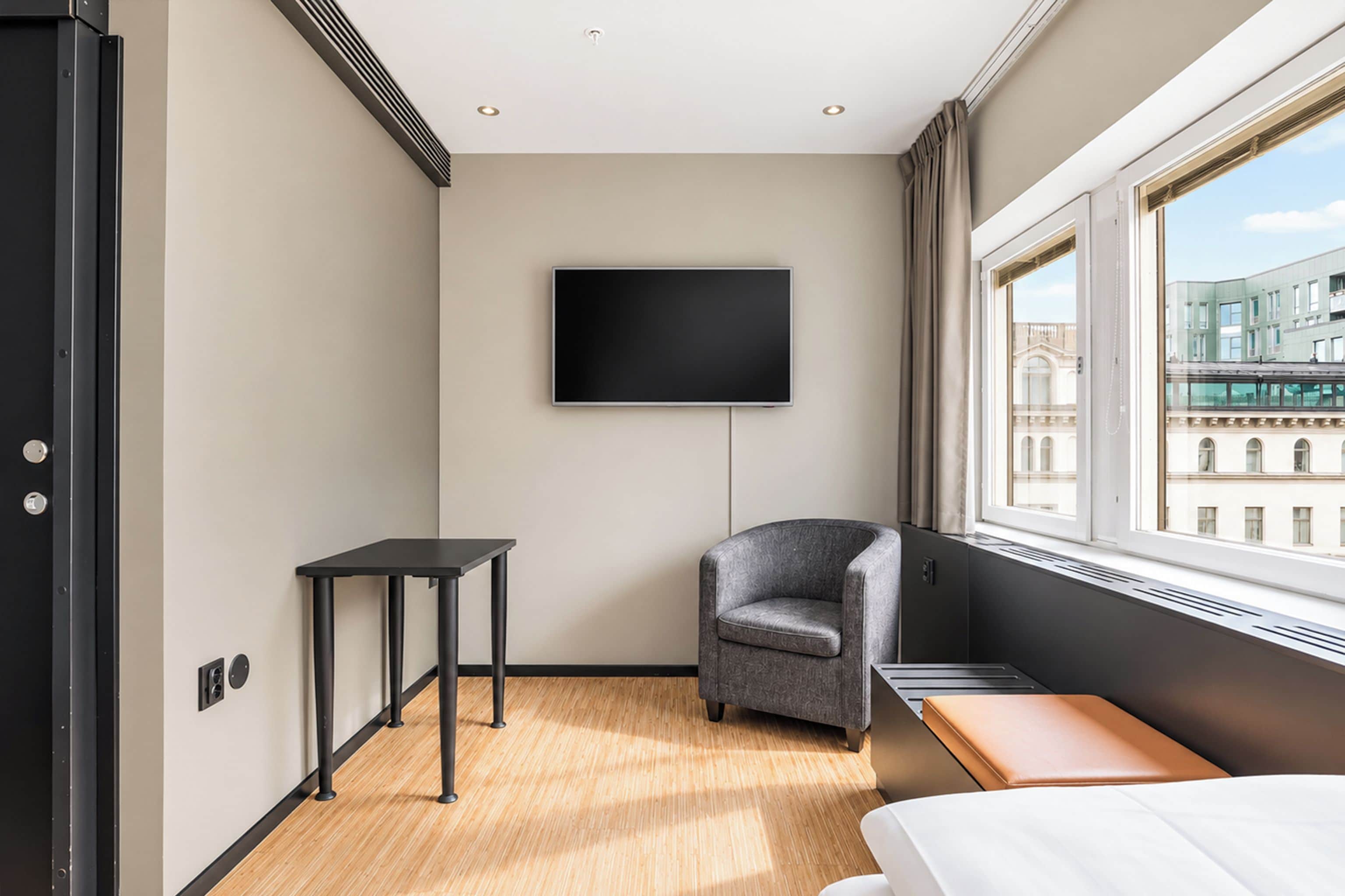 Habitación de hotel moderna con una televisión de pantalla plana montada en la pared, una mesita pequeña, un sillón gris, suelo de madera y un ventanal con vistas a la ciudad.
