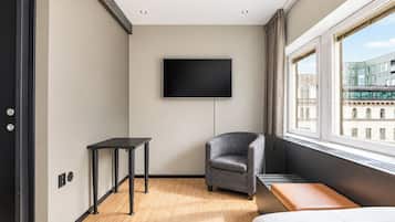 Habitación de hotel moderna con una televisión de pantalla plana montada en la pared, una mesita pequeña, un sillón gris, suelo de madera y un ventanal con vistas a la ciudad.
