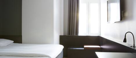 Una habitación de hotel moderna con una cama, un pequeño banco, un lavabo y una ventana con cortinas.
