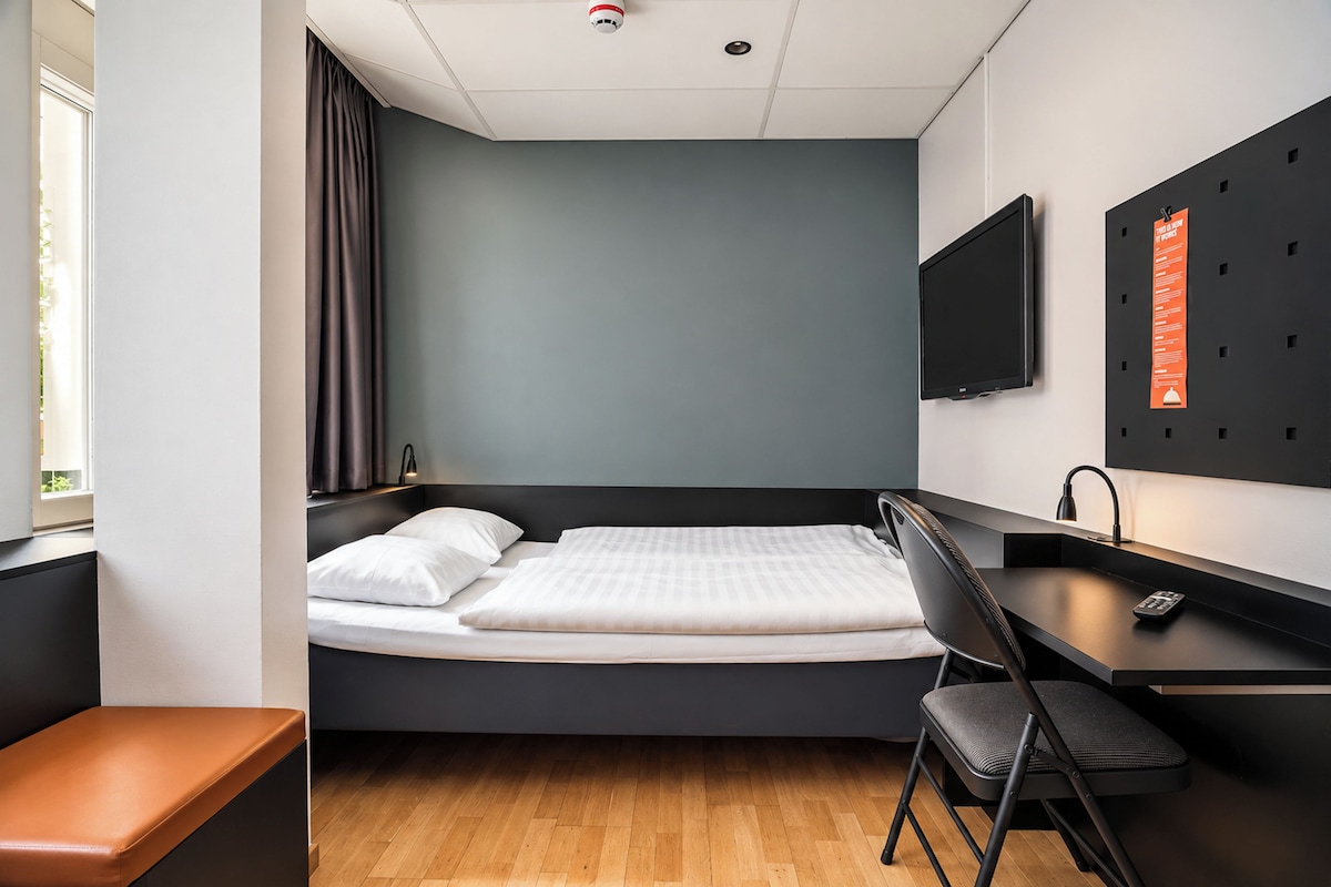 Een kleine hotelkamer met een bed, een bureau, een stoel, een televisie en een raam.