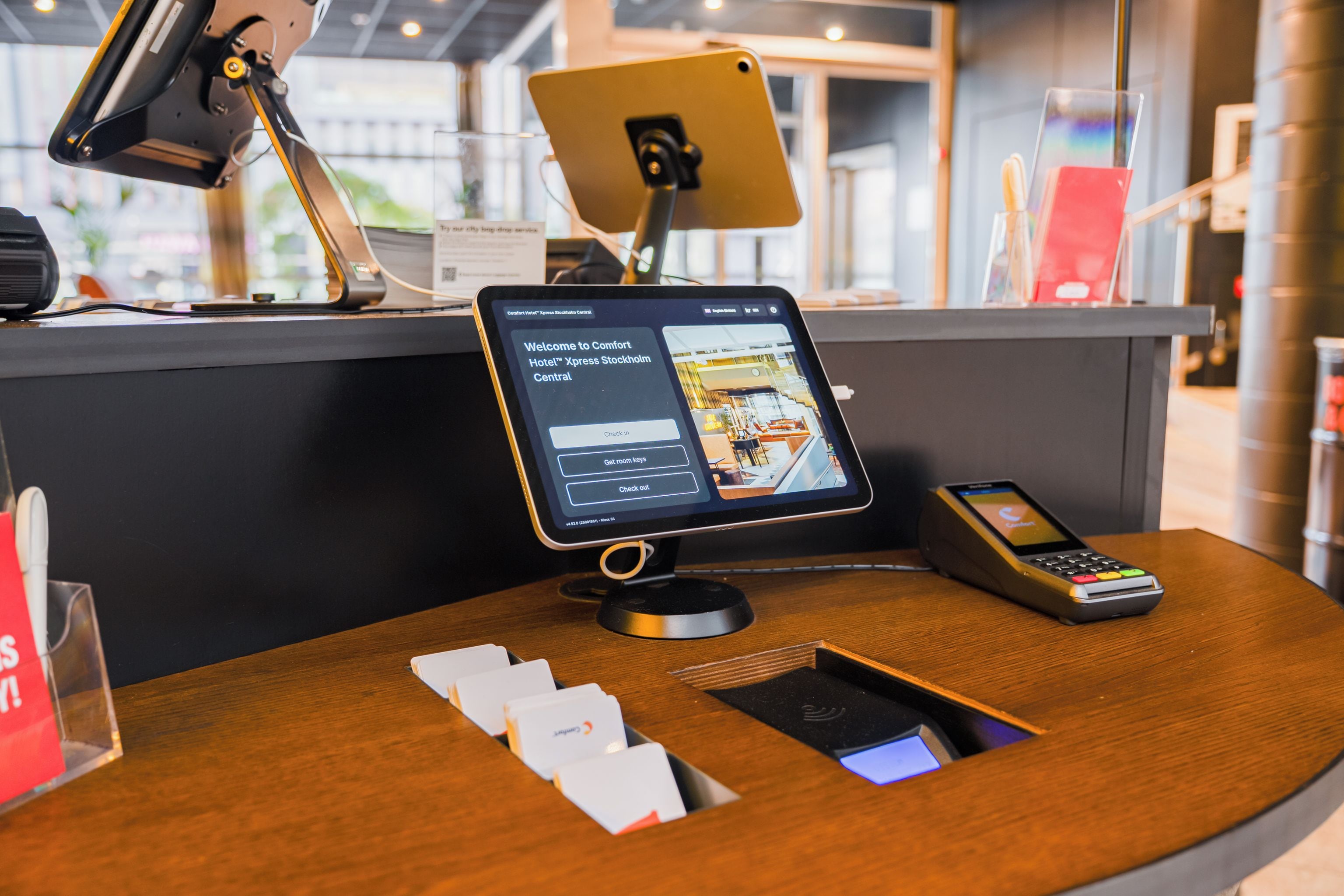 A hotel reception desk with a digital check-in kiosk, a mobile phone, and a tablet displaying the hotel's welcome screen.