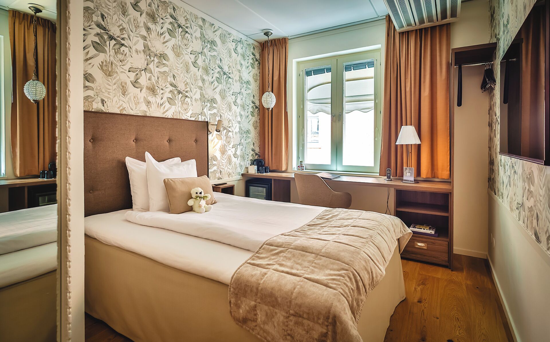 Ett hotellrum med en stor säng, ett skrivbord och en stol. Det finns ett fönster med gardiner, en lampa och en nallebjörn på sängen.
