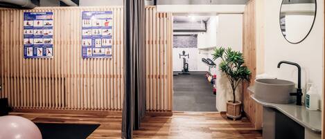 Une pièce moderne avec un plancher en bois, un tapis de sport noir, un ballon de gymnastique rose, un miroir fixé au mur, un lavabo et une plante.