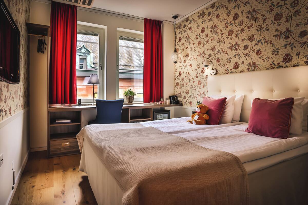 Ein Schlafzimmer mit einem Bett, einem Schreibtisch und einem Stuhl. Es gibt ein Fenster mit Vorhängen, eine Wand mit Blumenmuster-Tapete und eine Deckenleuchte.