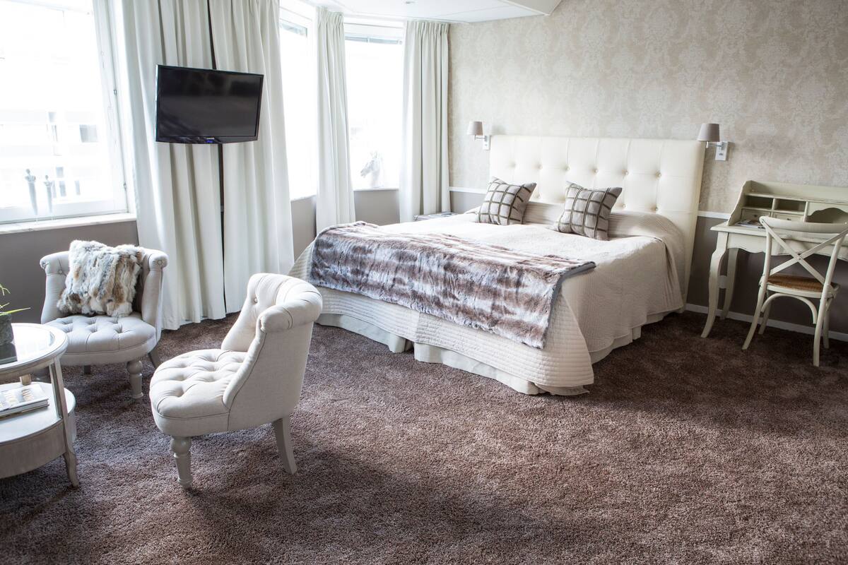 Ein Hotelzimmer mit einem großen Bett, einem an der Wand montierten Fernseher, einem Sessel, einem kleinen Tisch und einem Schreibtisch.