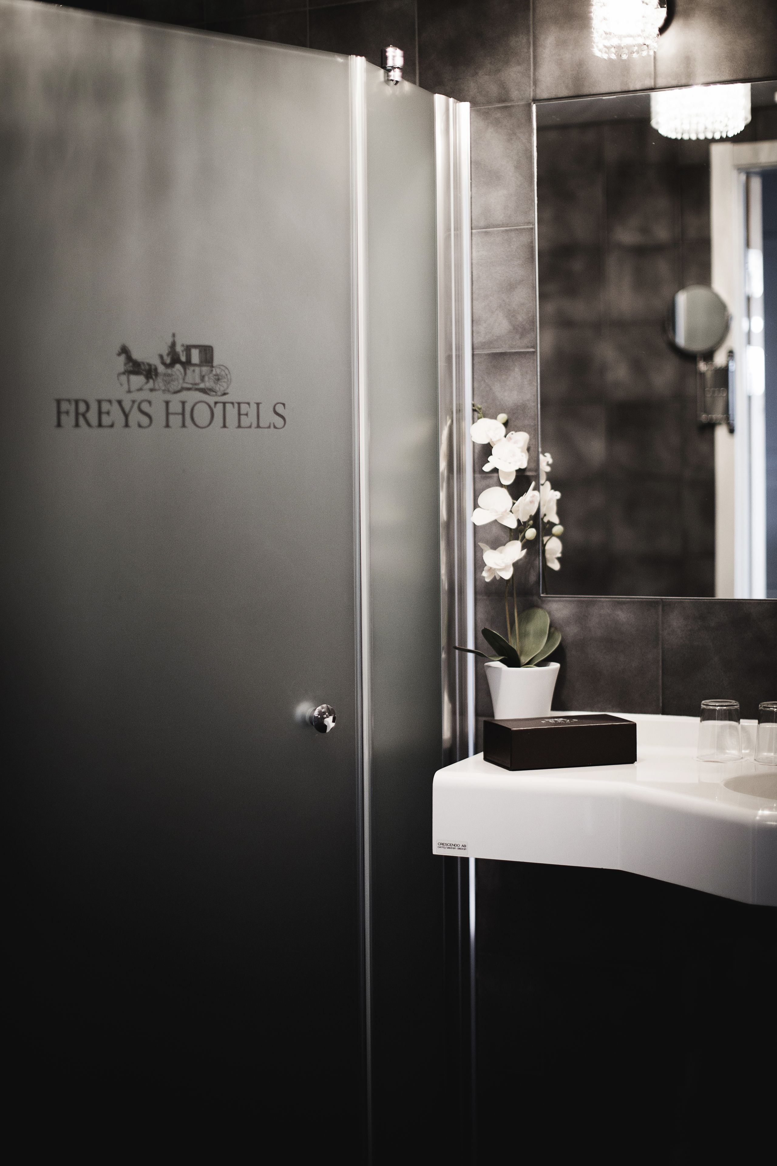 Un baño de Freys Hotels con un diseño moderno, que incluye un espejo grande, una orquídea en maceta y una zona de lavamanos elegante.
