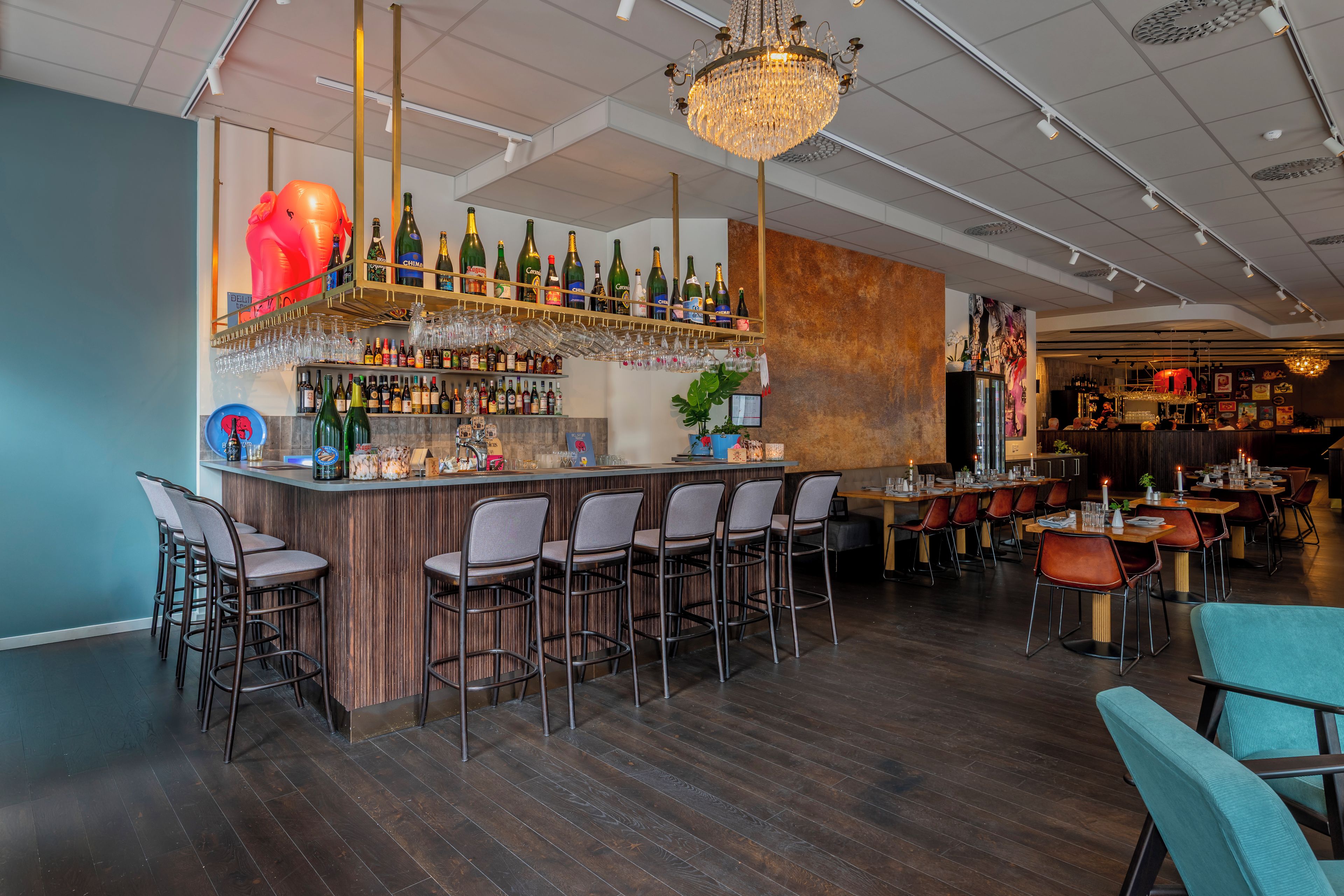 A bar area with a wooden counter, multiple bar stools, a chandelier, and a variety of bottles and glasses.