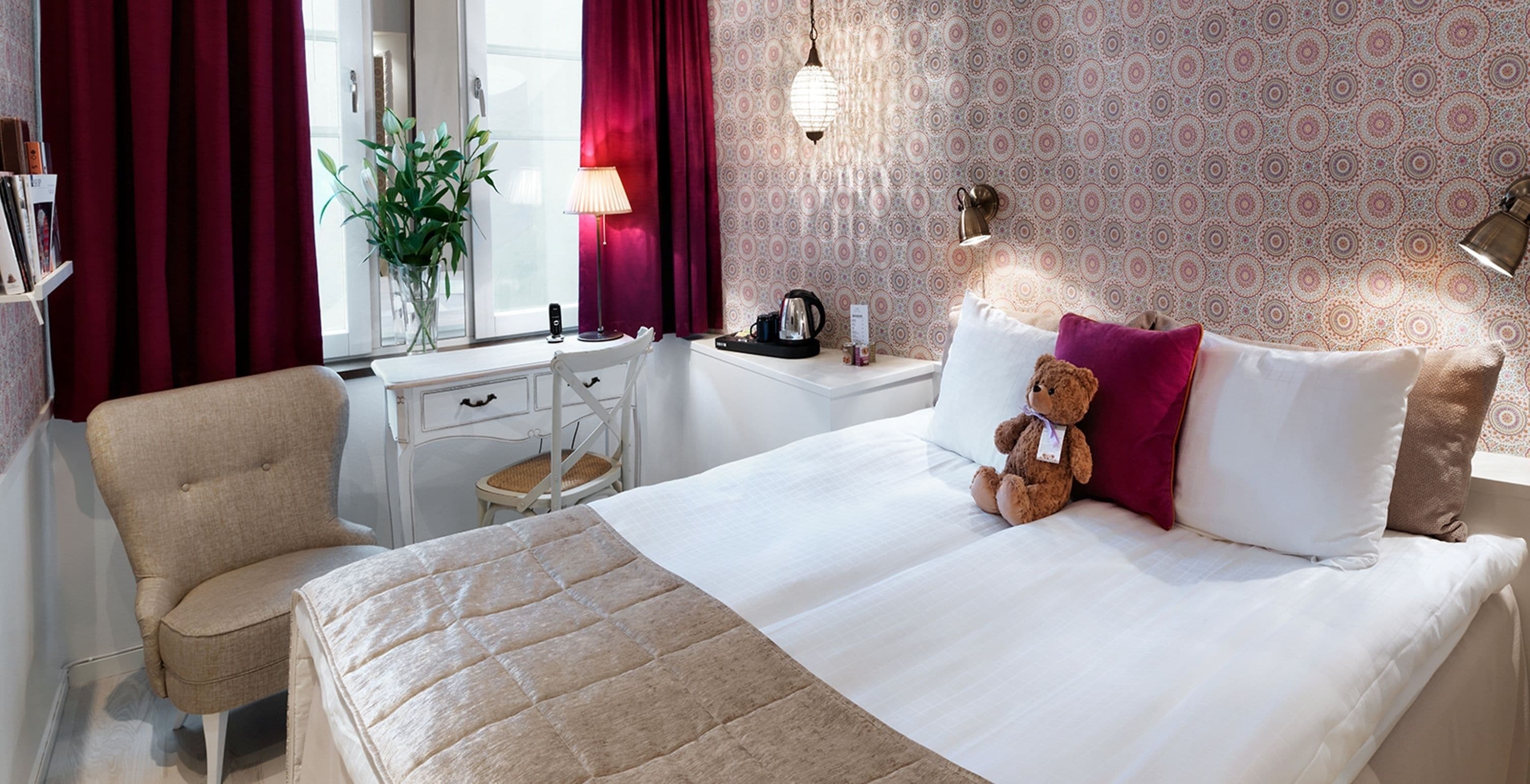 A hotel room with a bed, a chair, a desk, a lamp, and a teddy bear.