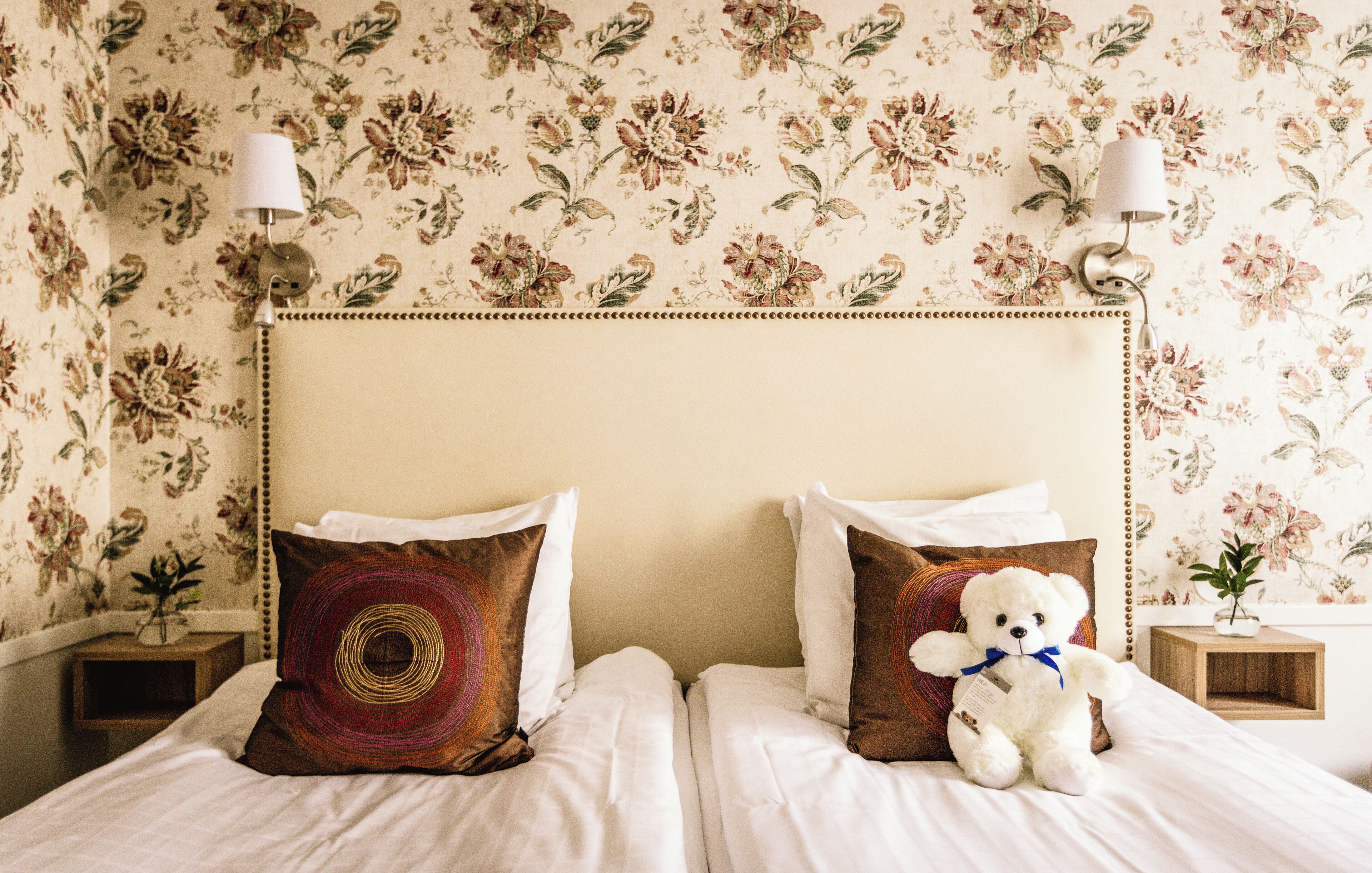 Una cama bien hecha con dos almohadas, un osito de peluche blanco y un estampado floral en el papel tapiz.