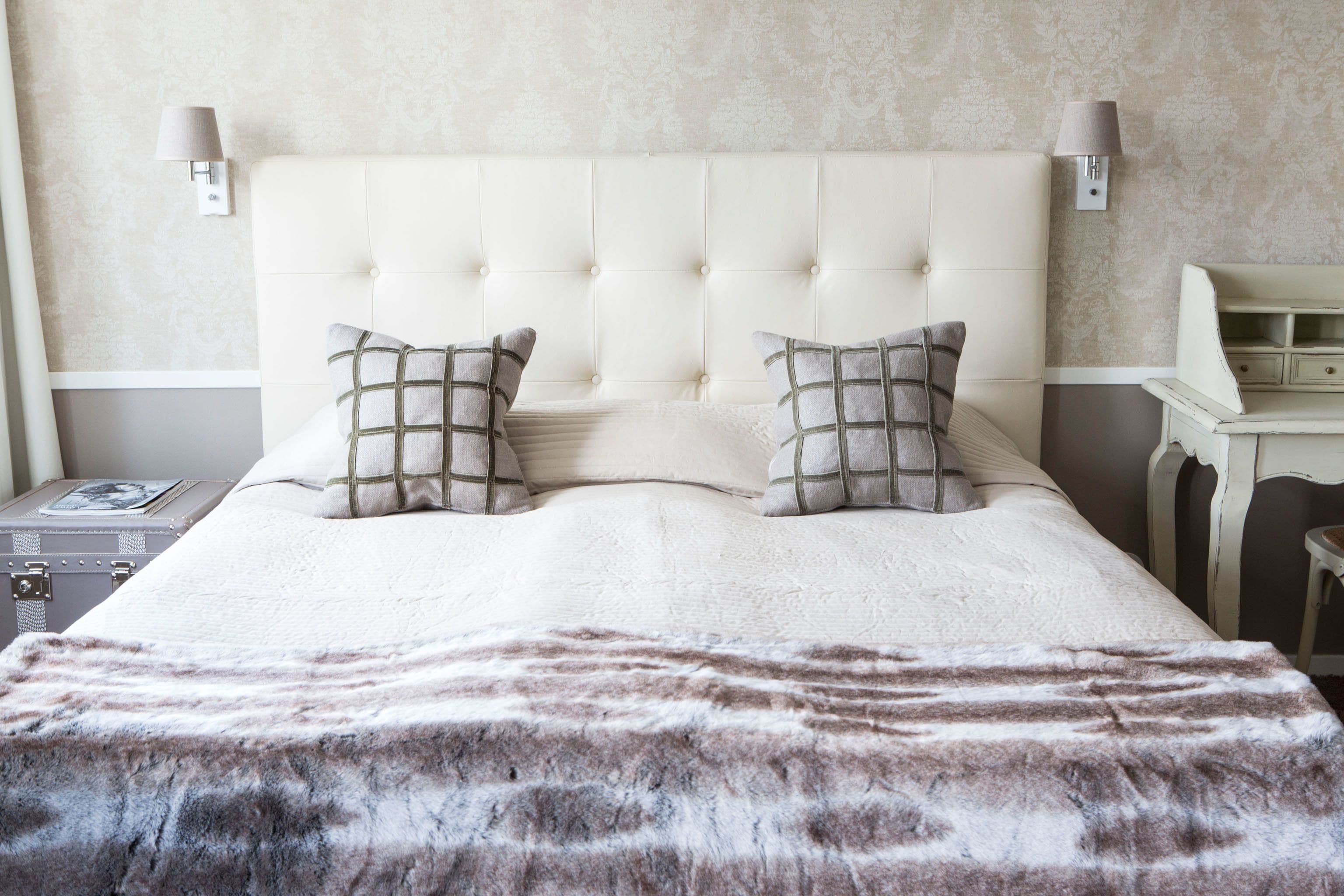 A neatly made bed with a tufted headboard, two bedside lamps, a nightstand with a lamp, and a suitcase on the left side.