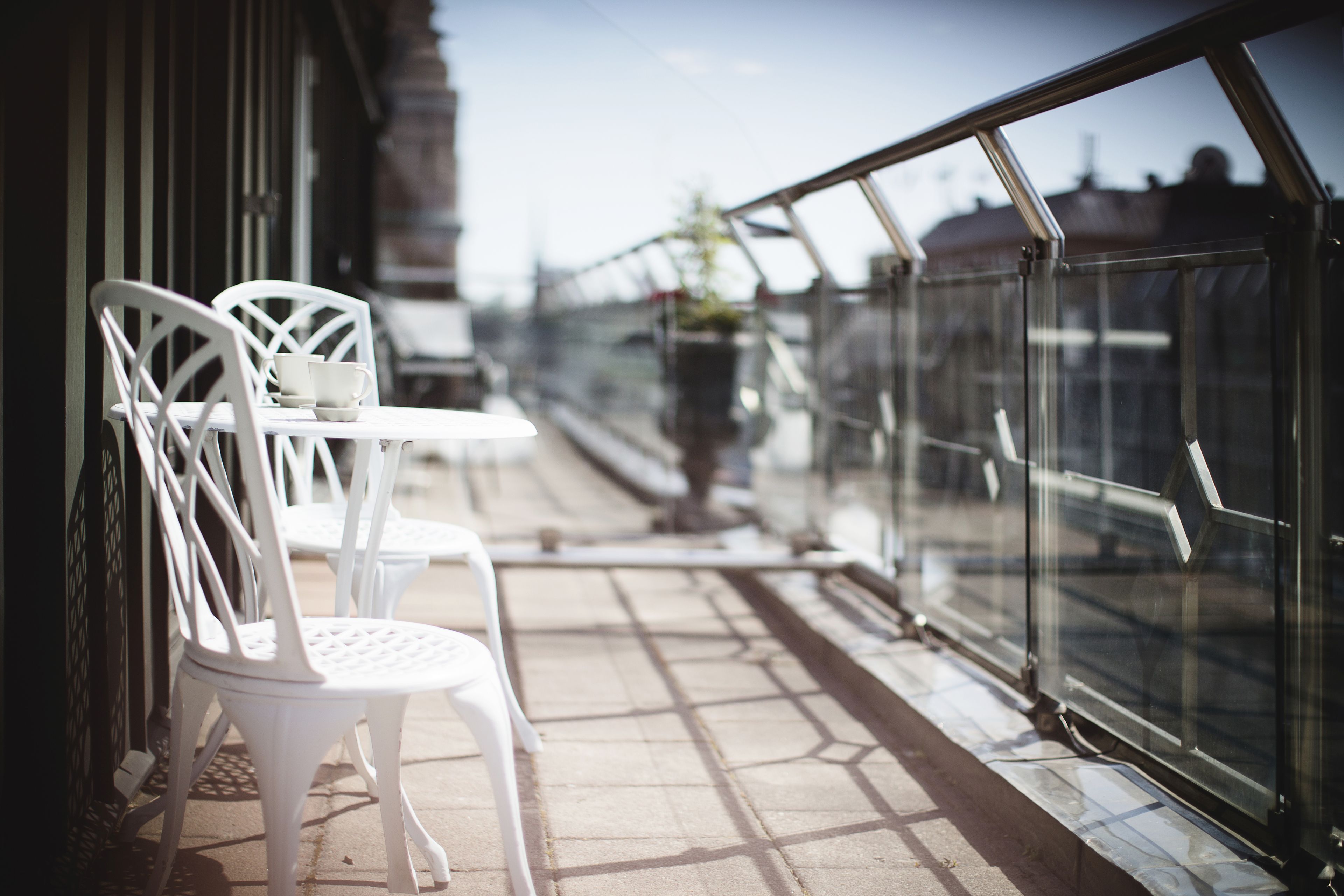 A balcony with white chairs and a table, overlooking a street with buildings and a clear sky.
