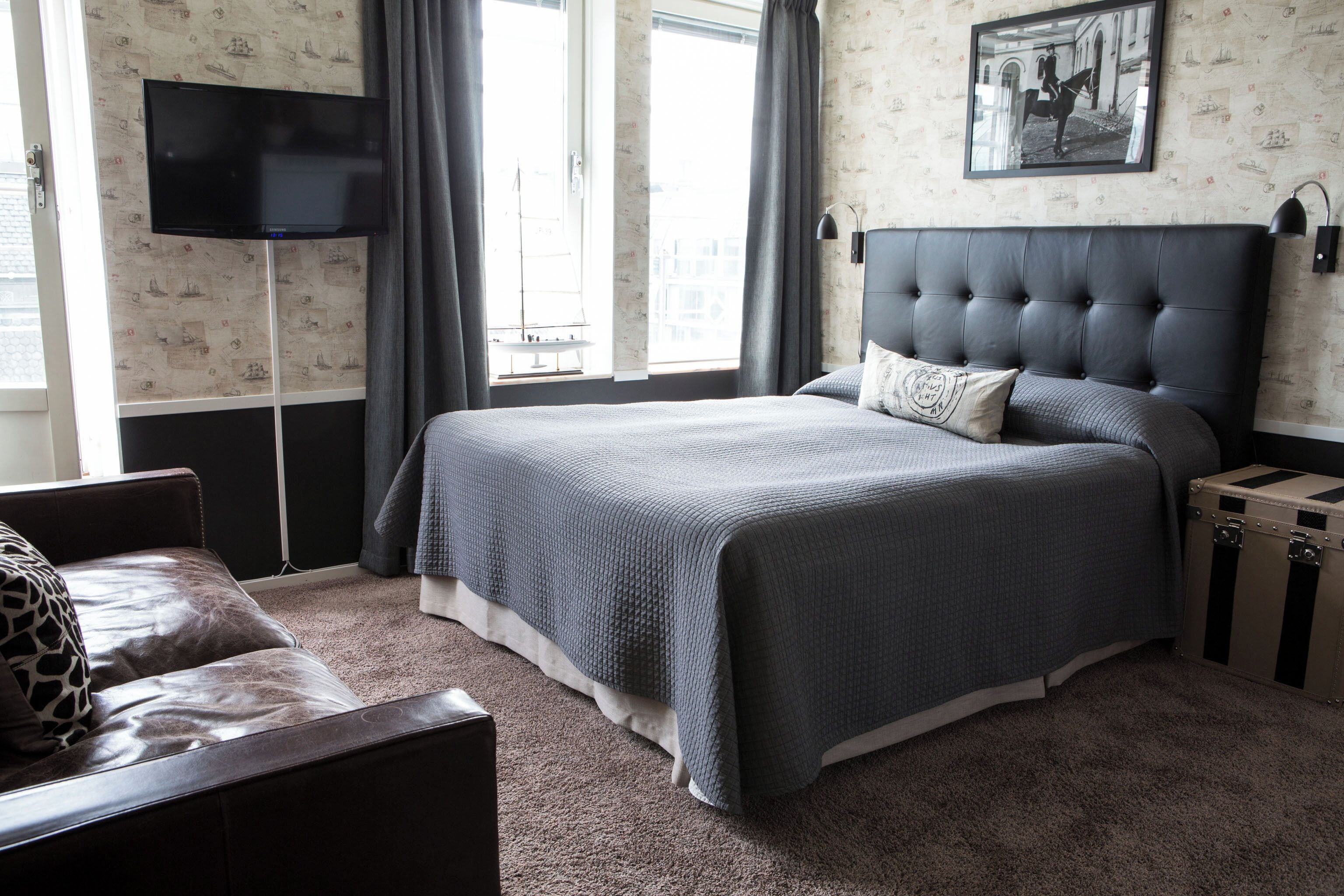 Une chambre avec un grand lit, un téléviseur à écran plat fixé au mur, une fenêtre avec des rideaux et une valise posée sur le sol.