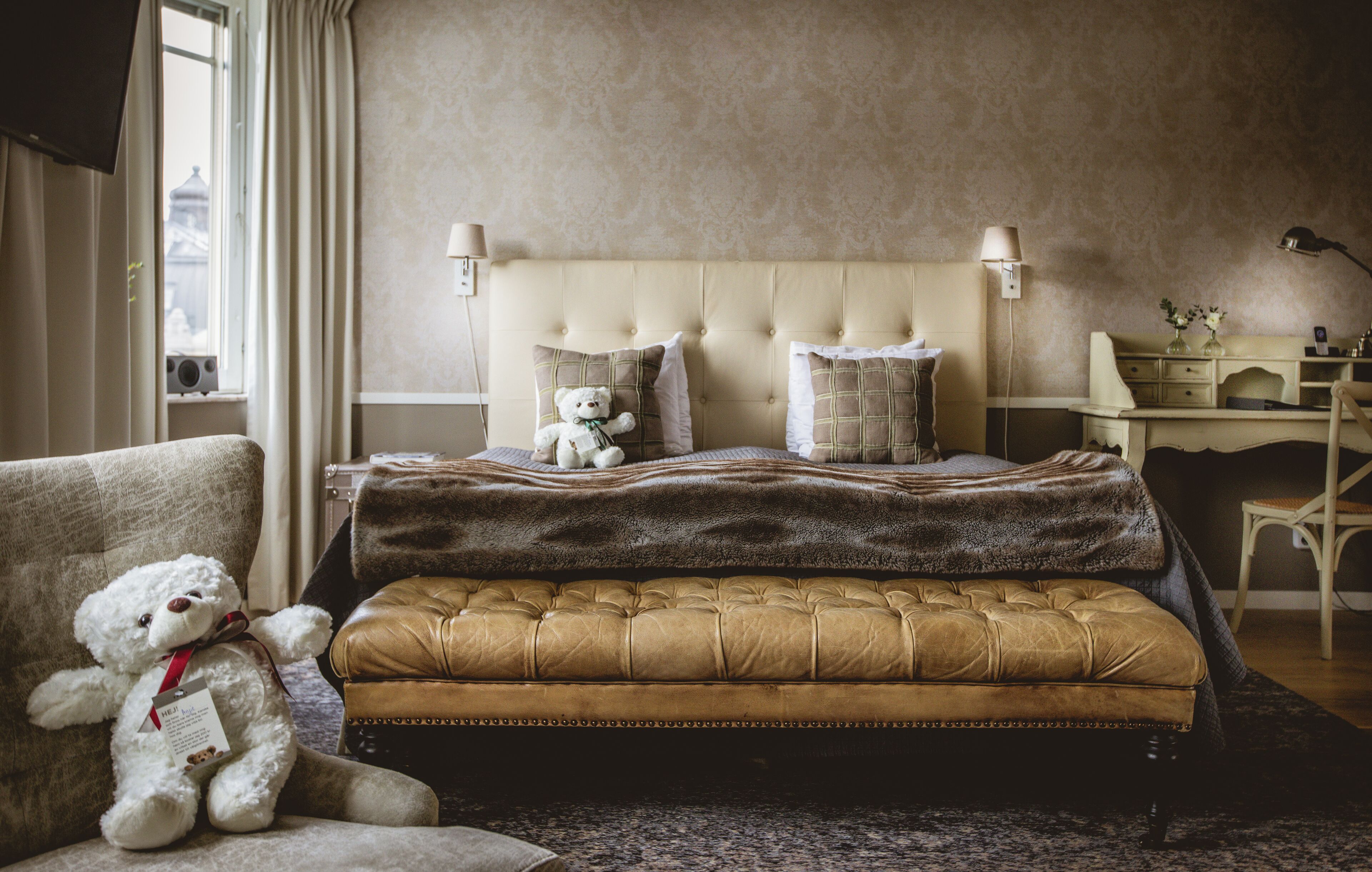 A bedroom with a tufted headboard, a bedside table, a lamp, a chair, and a teddy bear.