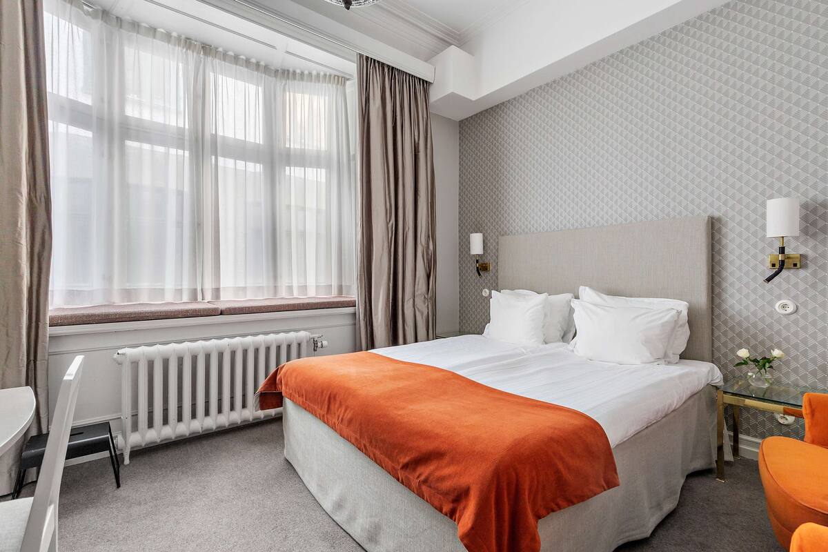 A hotel room with a bed, a desk, a chair, a radiator, and a window with curtains.