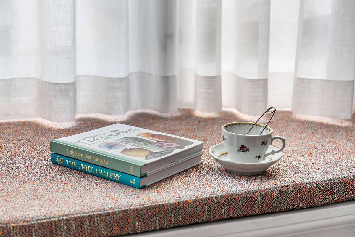 A stack of books on a textured surface near a window with sheer curtains.