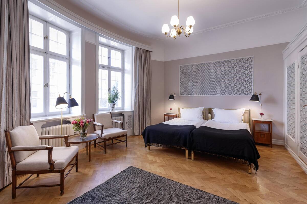 A well-lit bedroom with two beds, a sitting area with a chair and table, and large windows.